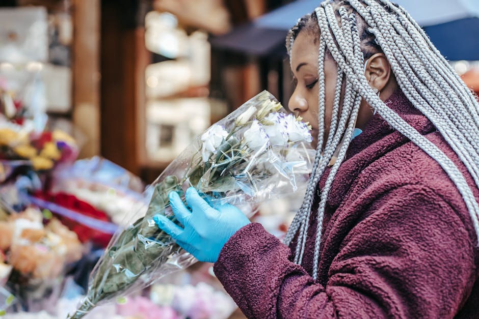 A woman with silver braided hair wearing a maroon fleece jacket and blue gloves is seen holding a bouquet of white roses wrapped in clear cellophane. She is smelling the flowers at an outdoor market s