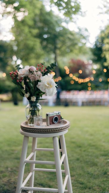 A glass vase filled with a fresh floral arrangement featuring white peonies, pink and white roses, and assorted greenery, placed on a white wooden stool outdoors. The bouquet is wrapped with clear cel