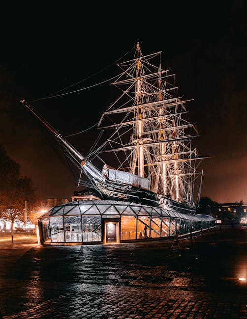 The image features the illuminated Cutty Sark ship at night, with its tall masts and rigging clearly visible against the dark sky. The ship is displayed on a modern, curved glass pavilion that serves
