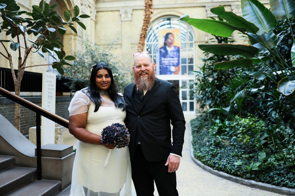 A bride and groom stand together indoors among lush green tropical plants and decorative foliage, with soft, natural lighting illuminating the scene. The bride has dark hair and medium skin tone, dres