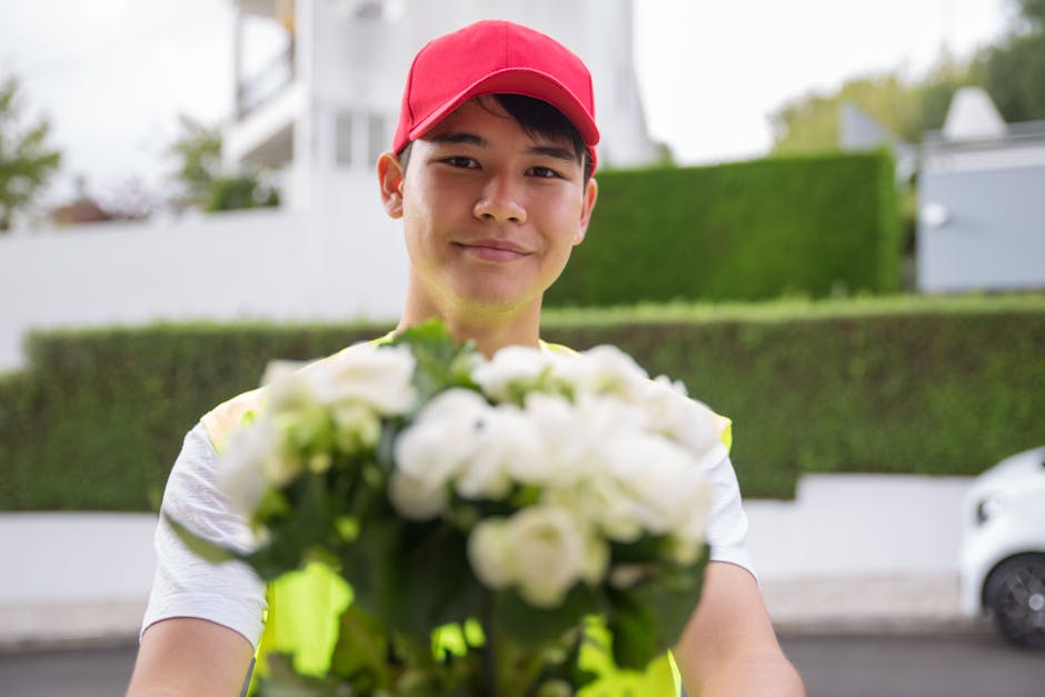 A delivery person from Greenwich Florist, dressed in a white shirt and a bright red cap, is standing outdoors holding a floral bouquet towards the camera. The bouquet features a collection of fresh wh