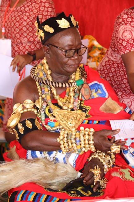 A person dressed in traditional Maasai attire, adorned with intricate gold jewelry including necklaces, bracelets, and rings, wearing a black and gold beaded headpiece. They are seated on a red fabric