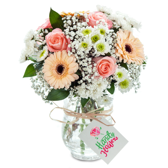 Set against a beautifully lit, crisp white background, this striking bouquet celebrates the serene charm of Greenwich. At its centre, fully opened roses in soft peach and blush tones captivate with their luxurious, spiraled petals-a tribute to the classic romance found in the area's elegant Georgian squares. Flanking these are two prominent gerbera daisies, their luminous peach-orange petals and contrasting dark eyes adding an exuberant splash akin to summer blooms along the Thames. Smaller white chrysanthemum blossoms with lime-green hearts are scattered throughout, juxtaposed with clouds of white gypsophila that enhance the bouquet's gentle volume and luminosity. Accents of deep green eucalyptus echo the freshness of local parks, while the artistry of the florist is clear in the thoughtfully layered textures and perfectly balanced silhouette. The entire arrangement is elegantly housed in a clear glass vase-its subtle pattern catching the light and allowing the stems' vivid emerald hues to shimmer in the water. A natural raffia ribbon binds the neck, from which a crisp white tag is suspended, ready for a hand-penned note. This display, prepared with talents honed in the heart of Greenwich, is ideal for occasions of gratitude, new beginnings, or drawing in a sense of peace and freshness to local homes from Blackheath to Maze Hill.