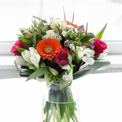 This exquisite bouquet, arranged with care near Greenwich's bustling Church Street, is a testament to balance and artistry. Four large gerbera daisies command attention: two ablaze in orange and two whispering in peach, their soft petals haloed around earthy brown centres reminiscent of autumn leaves fallen in Greenwich Park. Deep magenta roses, their petals lush and unfurling, weave romance through the heart of the arrangement, contrasting elegantly with clusters of snow-white alstroemeria and delicate sprigs of fill-perhaps the sort that thrive in shaded spots along the River Thames. Framing the blooms is a verdant wrap of broad aspidistra leaves, gracefully folded to echo the undulating greenery found on local riversides, all neatly gathered in a tall glass vase that reveals the tangle of fresh-cut stems. Diffused light sweeps across the bouquet, amplifying the flower colours without glare, casting reflections reminiscent of the sunlight that floods through Greenwich's historic markets. The bouquet's subtle blend of creamy tones, bright hues, and earthy greens evokes optimism, serenity, and celebration-making it ideal for gatherings, special occasions, or simply delivering a touch of heartfelt appreciation within the SE10 community. Every detail speaks to the practiced touch of a local florist, infusing Greenwich's distinctive charm into a timeless floral gift.